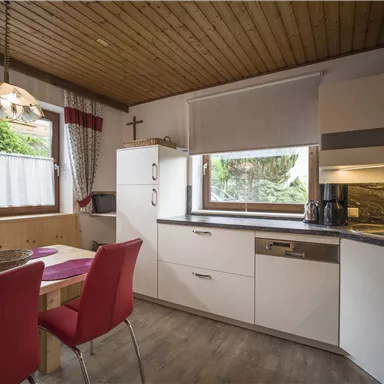 Eine moderne Küche mit weißen Möbeln und roten Stühlen. Große Fenster sorgen für viel Licht und einen Blick ins Freie.