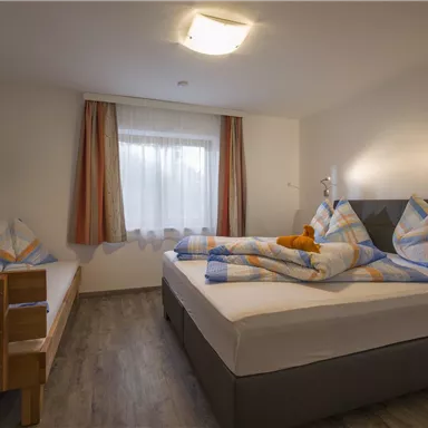 Ein modernes Schlafzimmer mit einem Doppelbett und einem Einzelbett. Es gibt ein Fenster mit Vorhängen und eine Wandmontage für einen Fernseher.