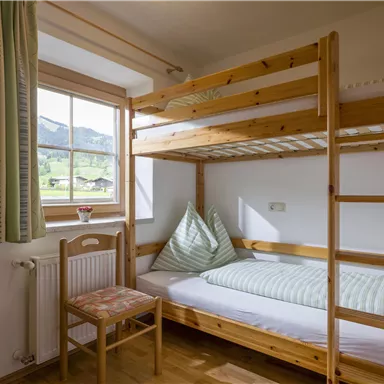 Ein gemütliches Zimmer mit einem Etagenbett aus Holz. Am Fenster sieht man eine schöne Aussicht auf die umliegenden Berge.