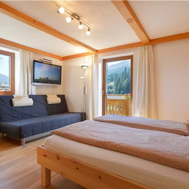 Ein gemütliches Zimmer mit zwei Betten und einem Schlafsofa. Große Fenster bieten einen Blick auf die Berge und viel Tageslicht.