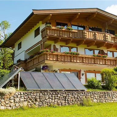 Ein gemütliches Haus im alpenländischen Stil mit einem großen Balkon. Umgeben von Grünflächen und einem sonnigen Himmel.