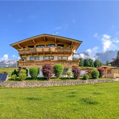 Ein wunderschönes Holzhaus in den Alpen mit großen Fenstern. Die Umgebung ist grün und mit Bäumen und Sträuchern gepflanzt.