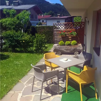 Ein gemütlicher Außenbereich mit einem Tisch und bunten Stühlen auf einem gepflegten Gartenweg. Im Hintergrund sind Pflanzen und eine Holzwand zu sehen, umgeben von einer schönen Berglandschaft.