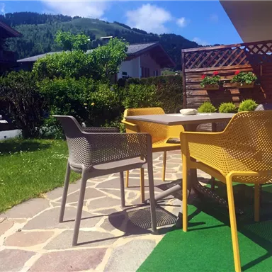 Ein gemütlicher Außenbereich mit einem Tisch und bunten Stühlen. Im Hintergrund sind grüne Sträucher und ein schöner Ausblick auf die Berge.