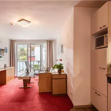 A cozy apartment with a red carpet and kitchenette. Large windows provide plenty of daylight and a view outside.