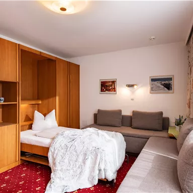 A cozy room with wooden cabinets and a modern sofa. There is a bed with white linens and bright windows that let in natural light.