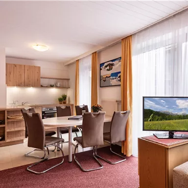 A bright and modern open-plan kitchen with a dining area and large windows. The room is cozily furnished and has a television.