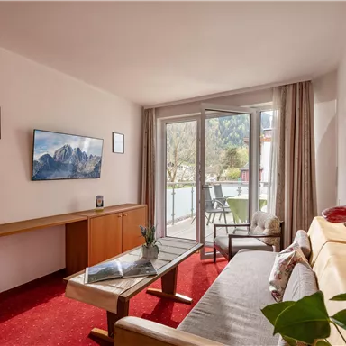 A modern living room with a sofa and a TV on the wall.  
Large windows let in a lot of light and show a view outside.