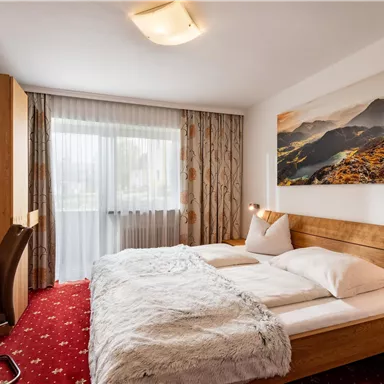 A cozy hotel room with two beds and a desk. Large windows let in plenty of light, and a beautiful landscape picture decorates the wall.