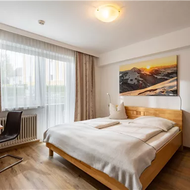 A cozy bedroom with a wooden bed and a large window. A picture of a mountain landscape hangs on the wall.