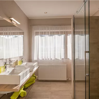 A modern bathroom with two sinks and large windows. The decor is bright and decorated with green towels.