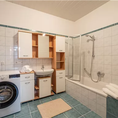 Ein modernes Badezimmer mit einer Badewanne und einer Dusche. Neben der Badewanne steht eine Waschmaschine und es sind frische Handtücher sichtbar.