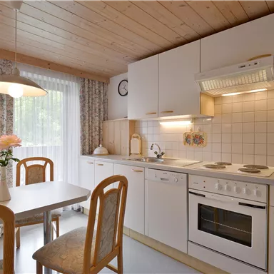 Eine freundliche Küche mit Holzdecke und hellen Möbeln. Der Tisch ist gedeckt und es gibt Fenster mit Vorhängen.