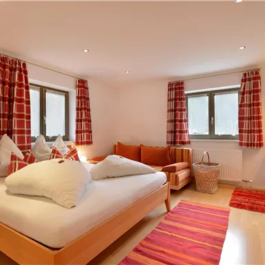 Ein helles Schlafzimmer mit einem großen Bett und einem gemütlichen Sofa. Rote Vorhänge und ein Teppich zusammen mit natürlichem Licht sorgen für eine einladende Atmosphäre.