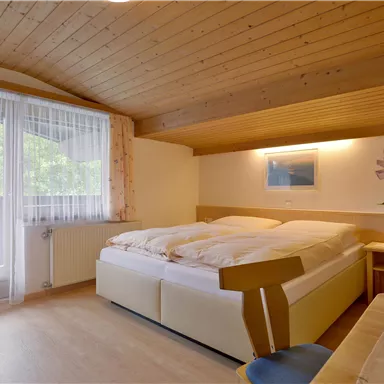 Ein gemütliches Schlafzimmer mit zwei Betten und Holzdecke. Das Zimmer hat ein Fenster mit Vorhängen und eine kleine Blumendekoration auf dem Tisch.