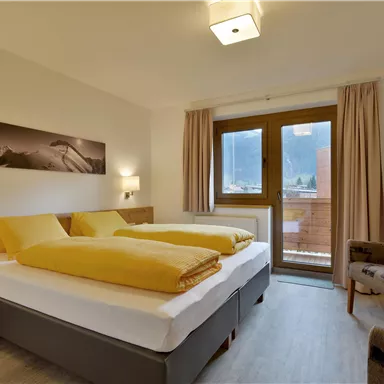 Ein modernes Schlafzimmer mit zwei Einzelbetten und gelben Decken. Große Fenster bieten Tageslicht und einen Blick nach draußen.