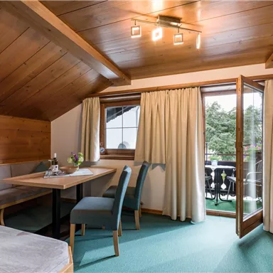 Ein gemütliches Zimmer mit Holzwänden und einem Balkon. Es gibt einen Esstisch und Stühle sowie große Fenster, die viel Licht hereinlassen.