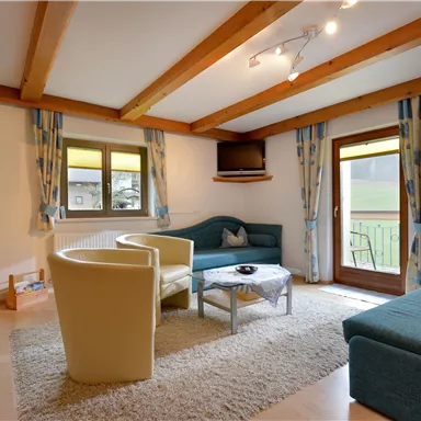 Ein gemütliches Wohnzimmer mit Holzbalken und modernen Möbeln. Es gibt ein Sofa, Sessel und einen Tisch vor einem Fenster mit Blick nach draußen.