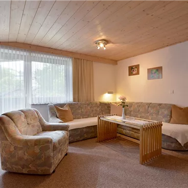 Ein gemütliches Wohnzimmer mit einem hellen Sofa und einem Sessel. Große Fenster sorgen für viel Tageslicht und eine einladende Atmosphäre.