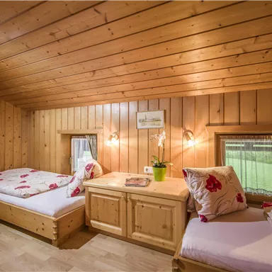 Ein gemütliches Schlafzimmer mit Holzvertäfelung und zwei Betten. Blumenmuster auf den Bettwäsche und ein Fenster mit Blick ins Grüne.