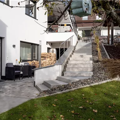 A modern house with a terrace and a paved path. A staircase leads to the upper area, surrounded by green areas and trees.
