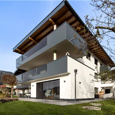 A modern residential building with several balconies and a clear, minimalist facade. The green lawn and the surrounding landscape create an inviting atmosphere.