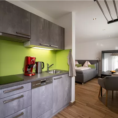 A modern kitchen unit with gray cabinets and green wall cladding. In the background, there is a cozy living area with a bed and an armchair visible.