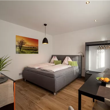 A modern bedroom with a double bed, a small seating area, and a table. The walls are brightly painted and a piece of artwork is mounted on the wall.