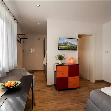 A modern room with bright curtains and a large window. There is a dining area, a TV on the wall, and a cozy seating area.