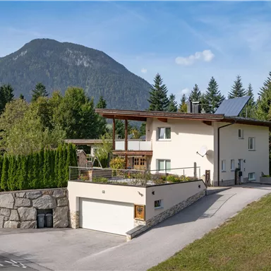 Ein modernes Haus mit einem begrünten Grundstück und einer eleganten Terrasse. Im Hintergrund sind Berge und Wälder sichtbar.