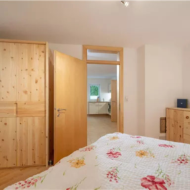 Ein gemütliches Schlafzimmer mit einem großen Bett und einer bunten Bettdecke. Der Raum ist hell und verfügt über Holzschränke sowie eine Tür zu einem angrenzenden Raum.