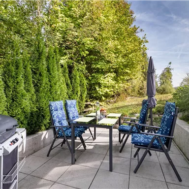 Ein gemütlicher Außenbereich mit einem Tisch, vier Stühlen und einem Grill. Umgeben von grünen Pflanzen und einem klaren Himmel.