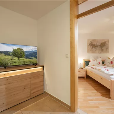 Ein modernes Wohnzimmer mit einem Flachbildfernseher und einem Holzschrank. Im Hintergrund ist ein einladendes Schlafzimmer mit einem Doppelbett und ansprechender Dekoration zu sehen.