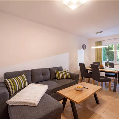 Ein modernes Wohnzimmer mit einer grauen Couch und einem Holztisch. Fenster mit grünen Vorhängen sorgen für viel Tageslicht.