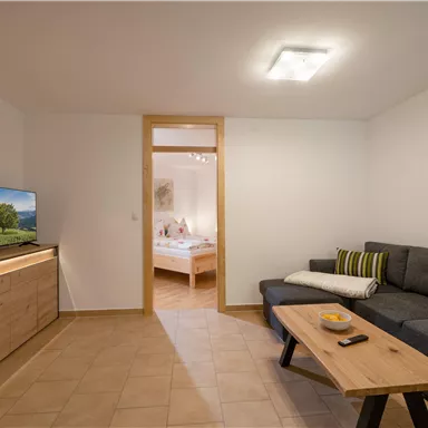 Ein gemütliches Wohnzimmer mit einem grauen Sofa und einem Holztisch. Im Hintergrund ist ein Schlafzimmer zu sehen, und ein Fernseher steht auf einem modernen Sideboard.