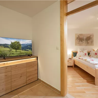 Ein gemütliches Schlafzimmer mit einem Doppelbett und sanften Farben. Auf der gegenüberliegenden Seite befindet sich ein Fernseher auf einem Holzmöbel.