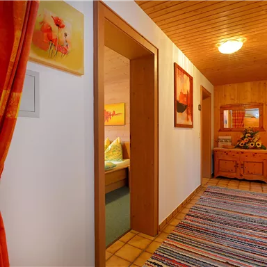 A bright hallway with wooden walls and colorful paintings. At the end of the hallway, there is a chest of drawers and a cozy room.