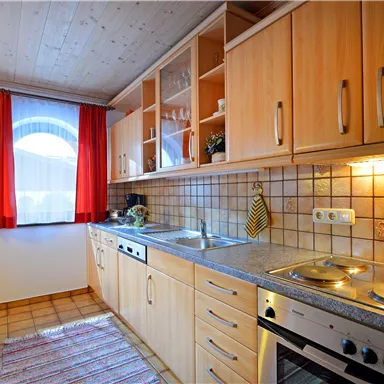 A cozy kitchen with wooden cabinets, a stove, and a sink. Red curtains and a window provide daylight.