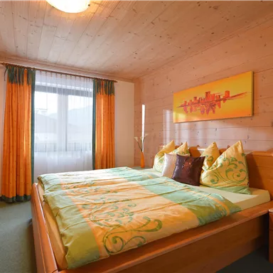 A cozy bedroom with wooden paneling and a large window. The bedding is in shades of green and yellow, and a modern artwork decorates the wall.