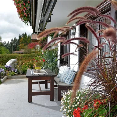 A cozy outdoor area with a wooden table and comfortable cushions. Colorful flowers and green grass surround the terrace.