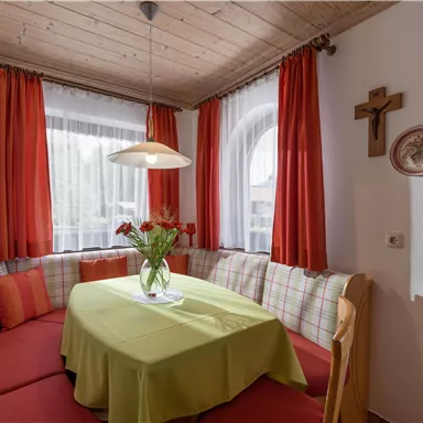 A cozy dining area with an L-shaped bench and red fabric. The table is decorated with a green tablecloth and a vase of flowers.