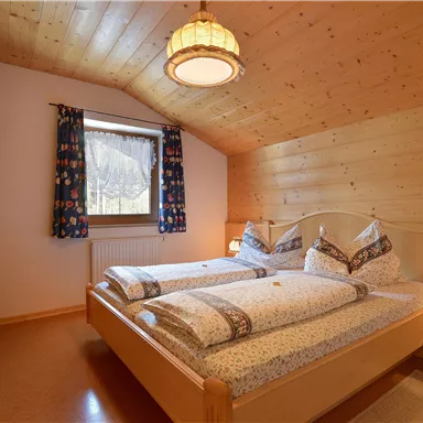 Ein gemütliches Schlafzimmer mit zwei Betten und Holzdecken. Große Fenster mit bunten Vorhängen lassen viel Licht herein.