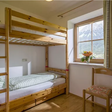 Ein einfaches Zimmer mit einem Etagenbett und einem Fenster, das einen Blick auf die Berge bietet. Auf dem Tisch steht eine Vase mit Blumen.