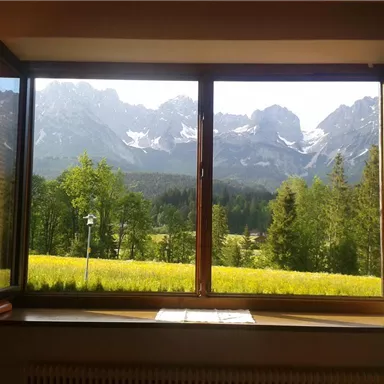 A view from a window of majestic mountains and lush forests. The landscape is adorned with green grass and blooming meadows.
