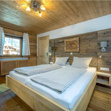 A cozy bedroom with a wooden bed frame and two pillows. The walls are clad in wood and a window provides daylight.