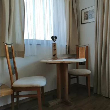 A small table with two chairs in a bright room. The window is covered with white curtains and there is a decorative vase on the table.