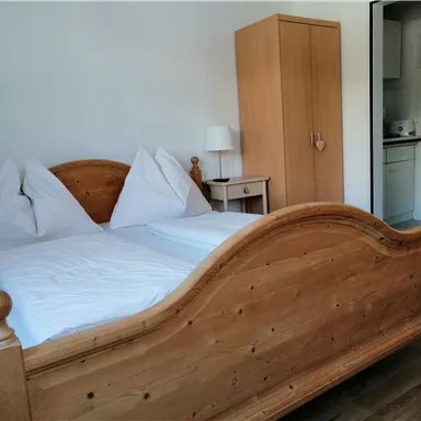 A cozy bedroom with a wooden bed and two pillows. There is a wardrobe against the wall and a bedside table with a lamp.