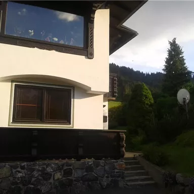 A beautiful house with a garden and a view of the mountains. The sky is partially cloudy, and there is a small path that leads to the garden.