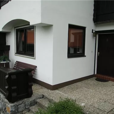 A house with a white facade and dark window frames. The terrace is equipped with a wooden railing and a bench.