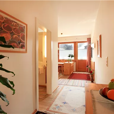 Ein heller Flur mit einer Pflanze und einem Tisch, auf dem Obst liegt. Im Hintergrund sind Fenster und ein Essbereich sichtbar.